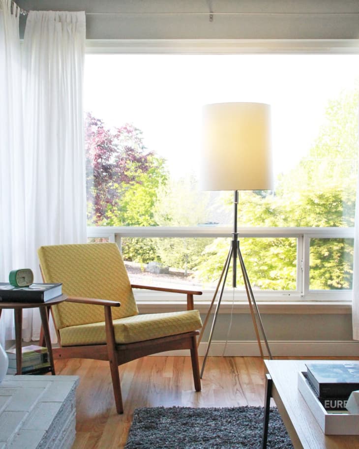 Mid-century modern living room with a yellow armchair, tripod floor lamp, and large window overlooking greenery.