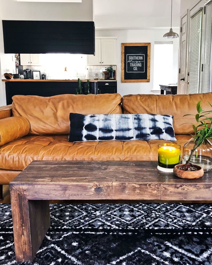 Brown leather sofa with a tie-dye pillow, wooden coffee table, green candle, and plant in a modern living room.