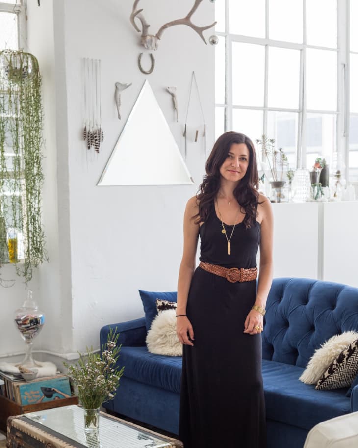 Woman in black dress standing in a living room with blue sofa, antler decor, and plants.