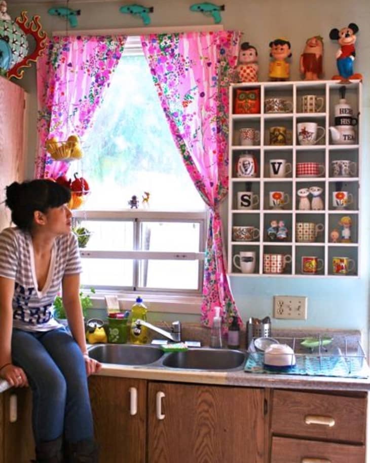 Woman sitting on kitchen counter with pink floral curtains, colorful figurines, and a wall shelf displaying mugs and decor.