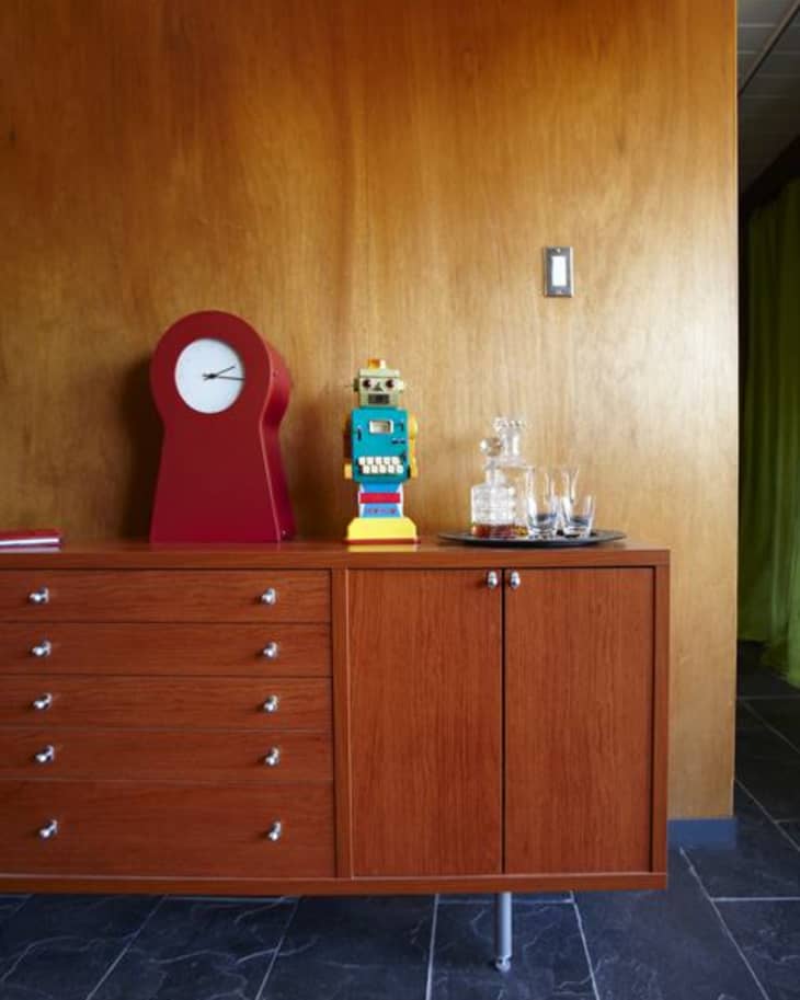 Mid-century modern sideboard with red clock, vintage robot toy, and glass decanter set against a wooden wall.
