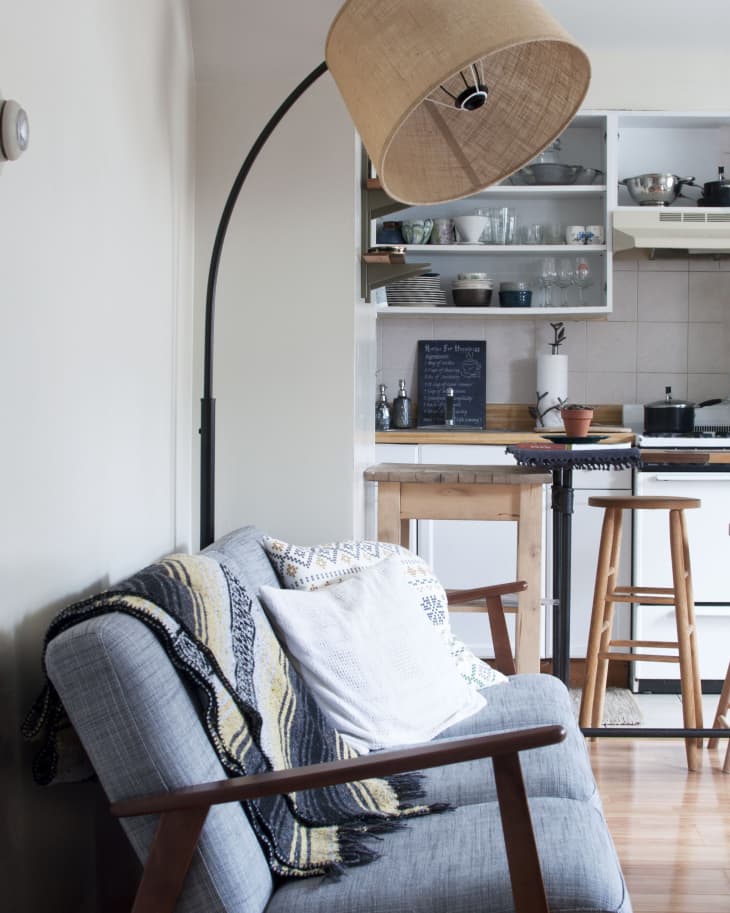Cozy living room with a gray sofa, striped throw, floor lamp, and view into a kitchen with wooden stools and open shelves.