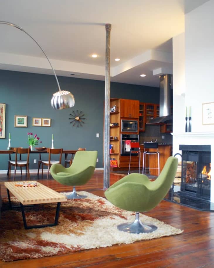 Open-concept living room with green swivel chairs, wooden dining table, modern fireplace, and kitchen with wood cabinets.
