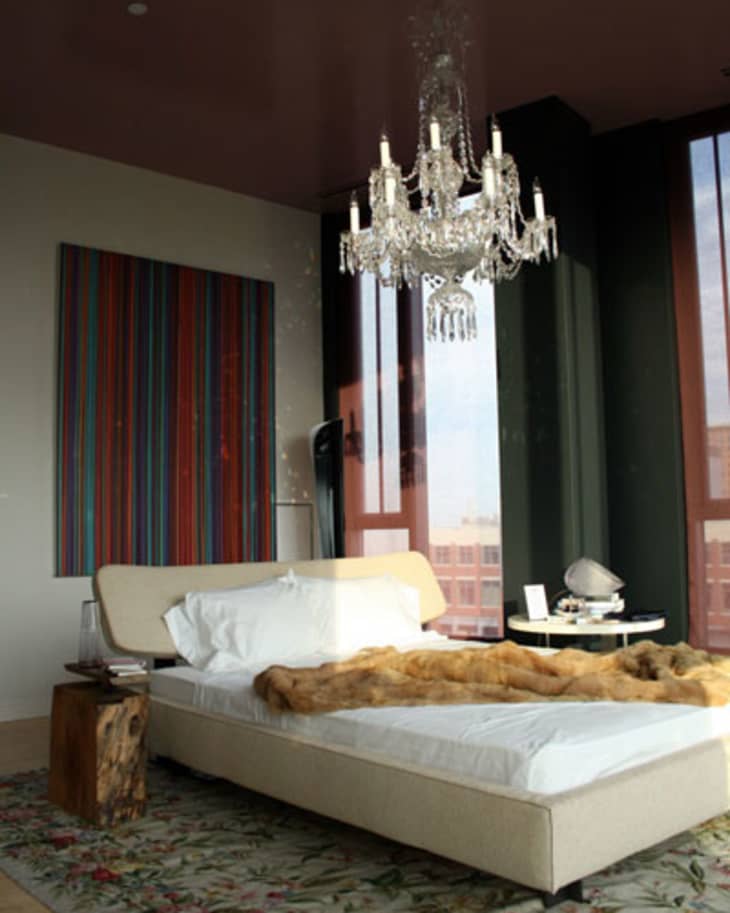 Modern bedroom with a chandelier, beige bed, fur throw, colorful wall art, and large windows.