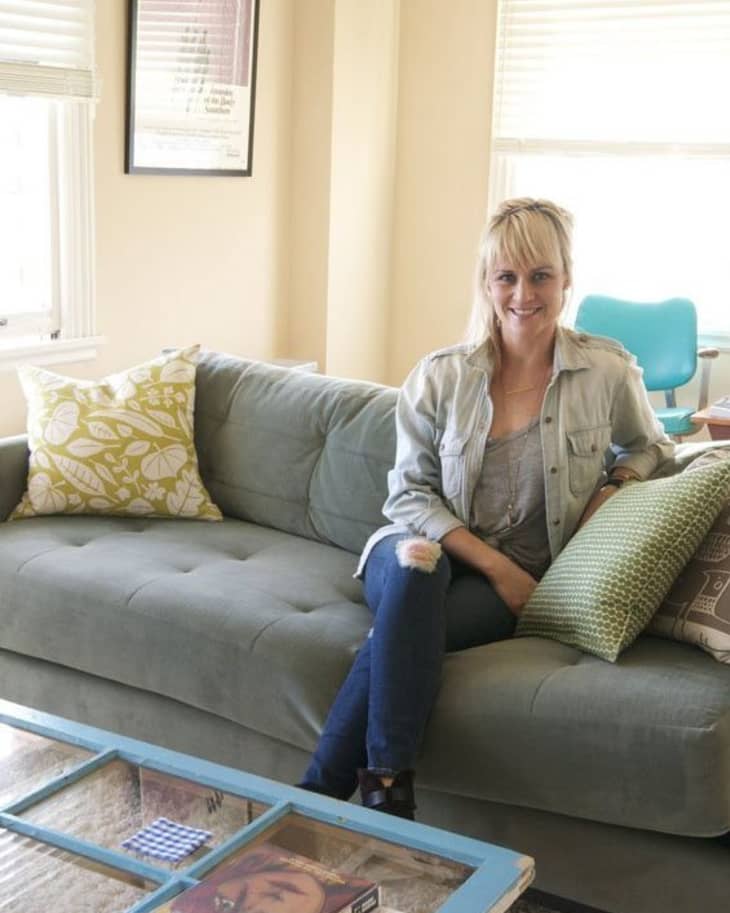 Woman sitting on a gray sofa with patterned pillows, next to a glass coffee table with books, in a bright living room.