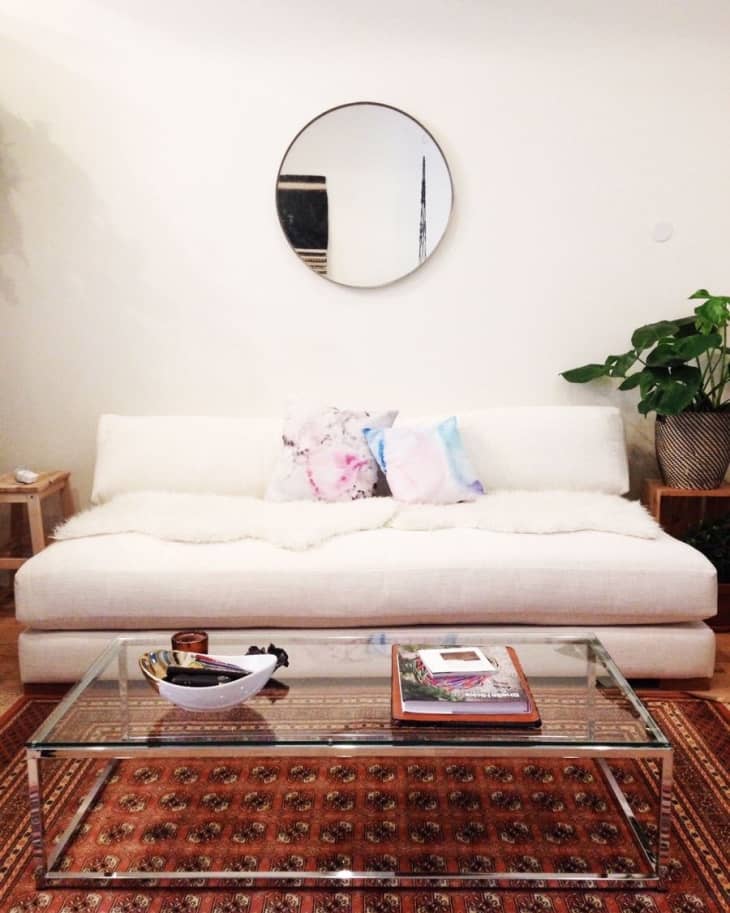 Cream sofa with colorful pillows, glass coffee table, round mirror, and potted plant on patterned rug.