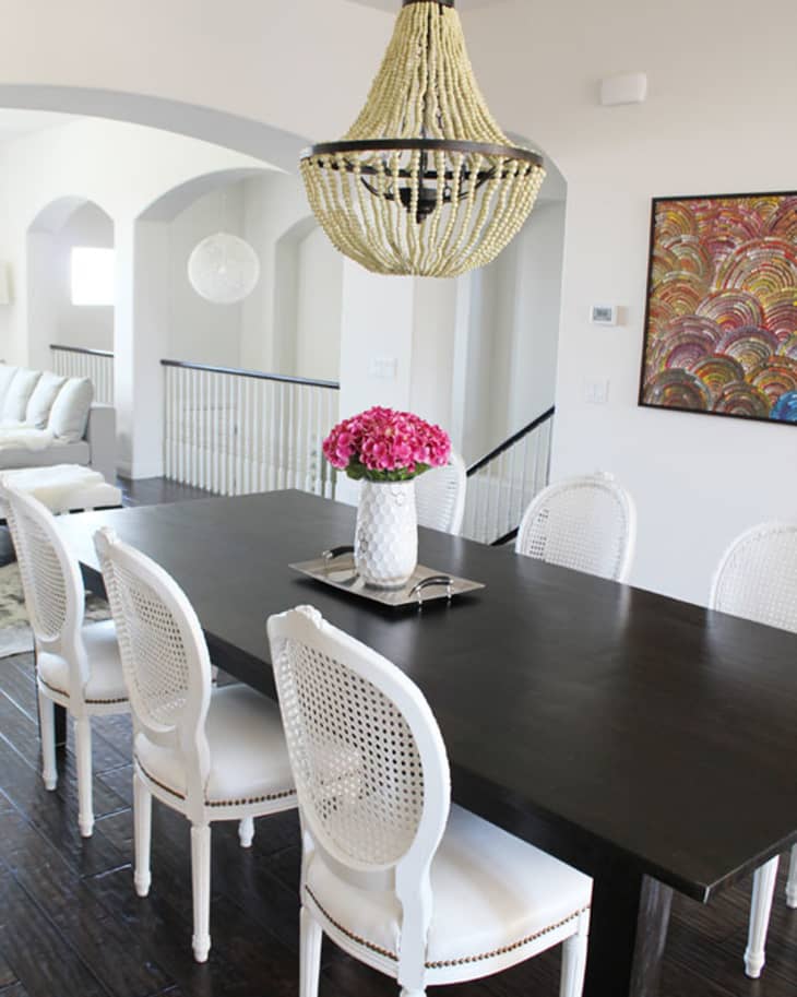Dining room with dark wood table, white chairs, pink flowers in vase, beaded chandelier, and colorful wall art.