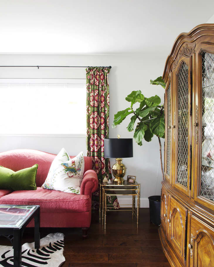 Red sofa with green and floral pillows, gold lamp on side table, patterned curtains, and wooden cabinet in living room.