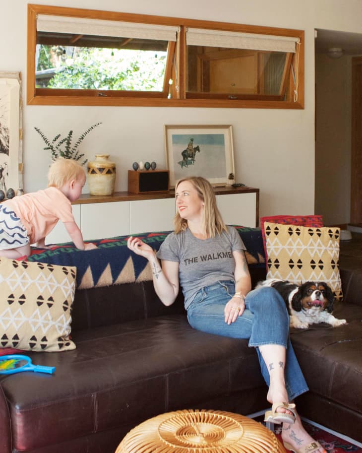 Woman sitting on a dark sofa with a child climbing and a dog resting, surrounded by patterned pillows and decor.