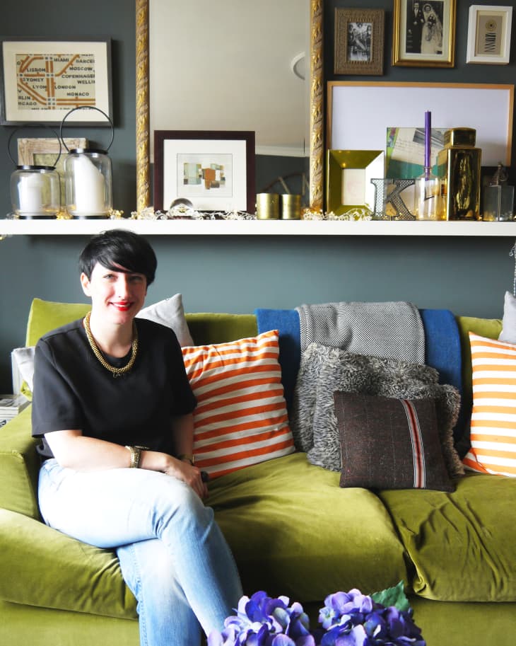 Woman sitting on green sofa with striped pillows, surrounded by eclectic wall art and decor, purple flowers in foreground.