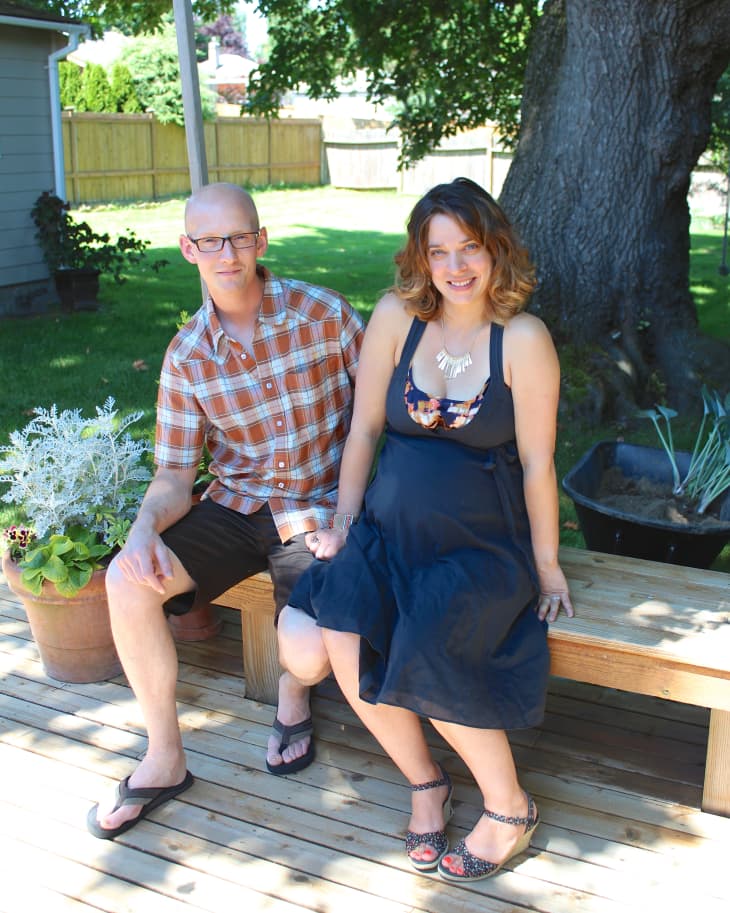 Man and woman sitting on a wooden deck bench, surrounded by potted plants, with a large tree in the background.