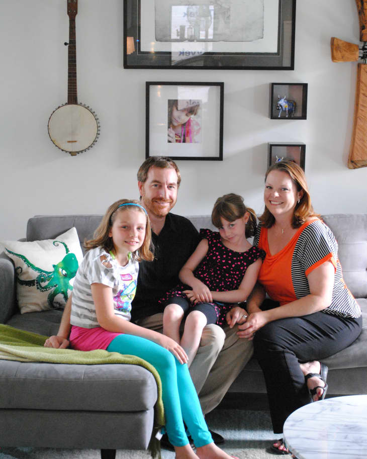 Family sitting on a gray sofa in a living room with framed photos, a banjo, and a marble coffee table.
