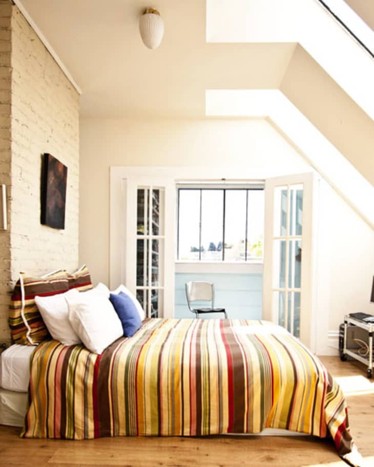 Loft bedroom with striped bedding, skylight, and French doors leading to a small balcony with a chair and computer desk.