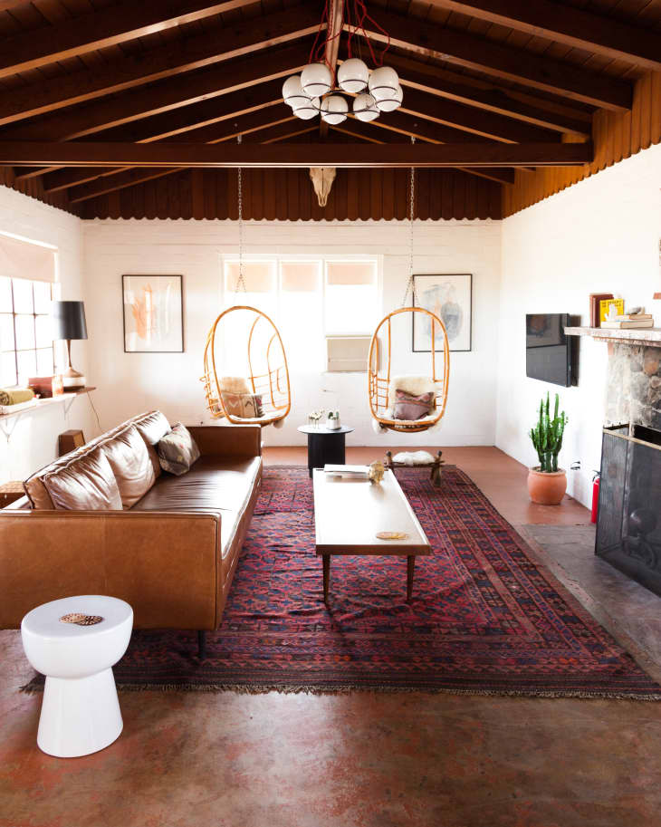 Mid-century living room with leather sofa, hanging chairs, red patterned rug, and wooden ceiling beams.