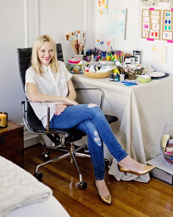 Woman in a white shirt and jeans sitting on a black office chair in a colorful art studio with supplies and artwork.