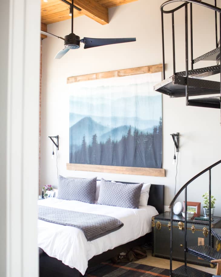 Loft bedroom with a spiral staircase, ceiling fan, mountain artwork, and a bed with gray pillows and blanket.