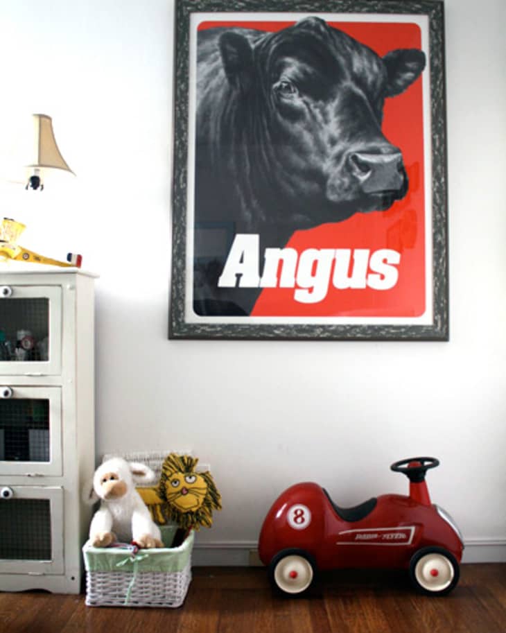 Framed Angus cow poster above a toy basket and red ride-on car in a child's room.