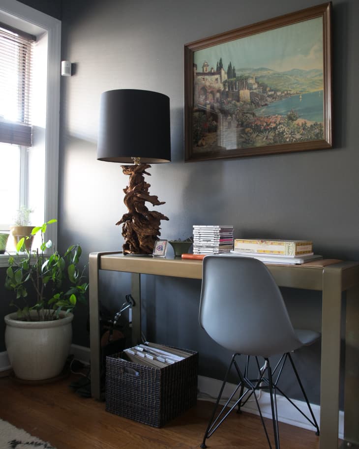 Home office with a gray chair, wooden desk, unique lamp, framed landscape art, and a potted plant by the window.