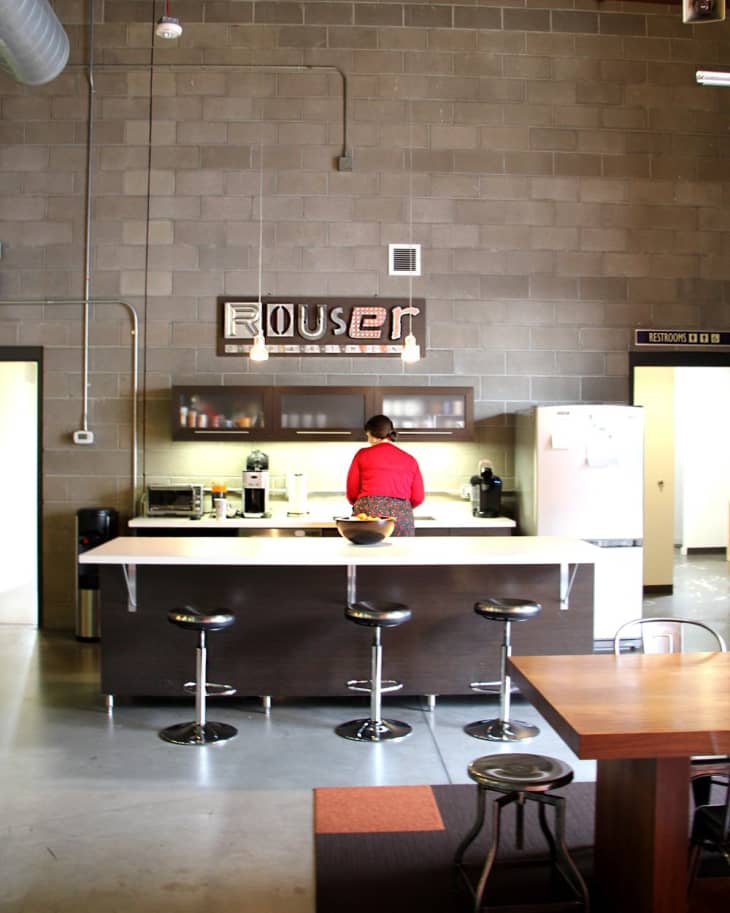 Industrial-style kitchen with bar stools, a person in red, and a "Rouser" sign on the wall.