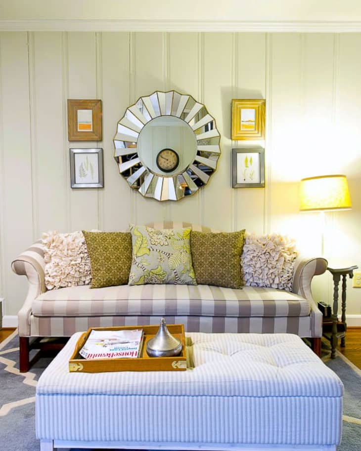 Striped sofa with decorative pillows, round mirror, framed art, and a lamp. Ottoman with tray and magazine on a patterned rug.