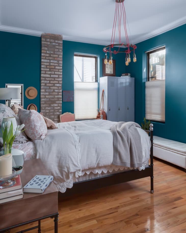 Bedroom with teal walls, brick accent, white bedding, pink floral pillows, and a red rope chandelier.