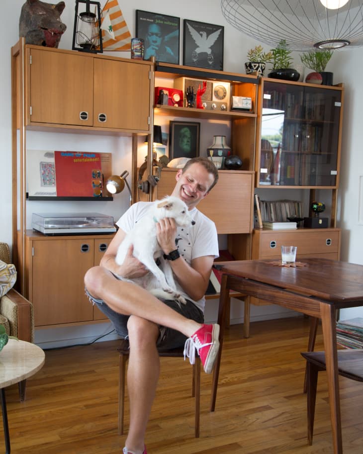 Man sitting in a mid-century modern living room, holding a white dog, with wooden shelves and eclectic decor in the background.