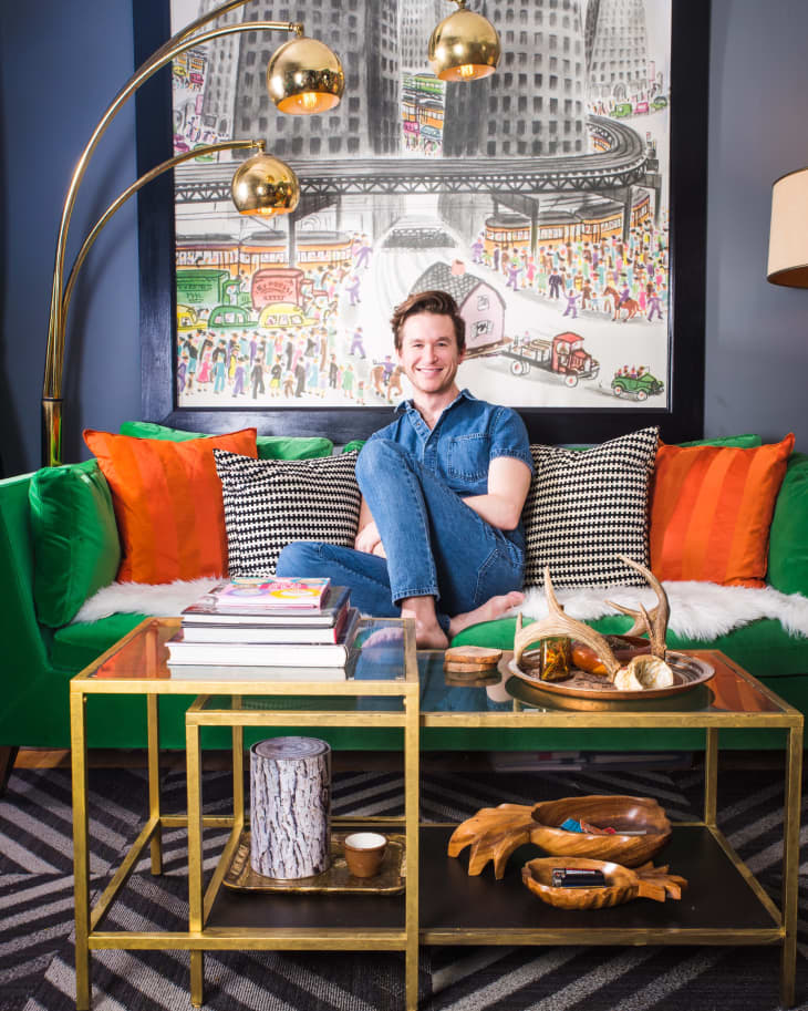 Man sitting on a green sofa with colorful cushions, under a gold arc lamp, in front of a cityscape artwork.