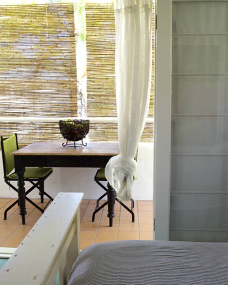 Small patio with a wooden table, two folding chairs, potted plant, bamboo blinds, and white curtain.