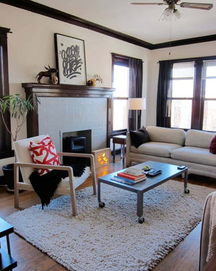 Living room with a beige sofa, armchair with red pillow, coffee table, fireplace, and large windows with dark curtains.