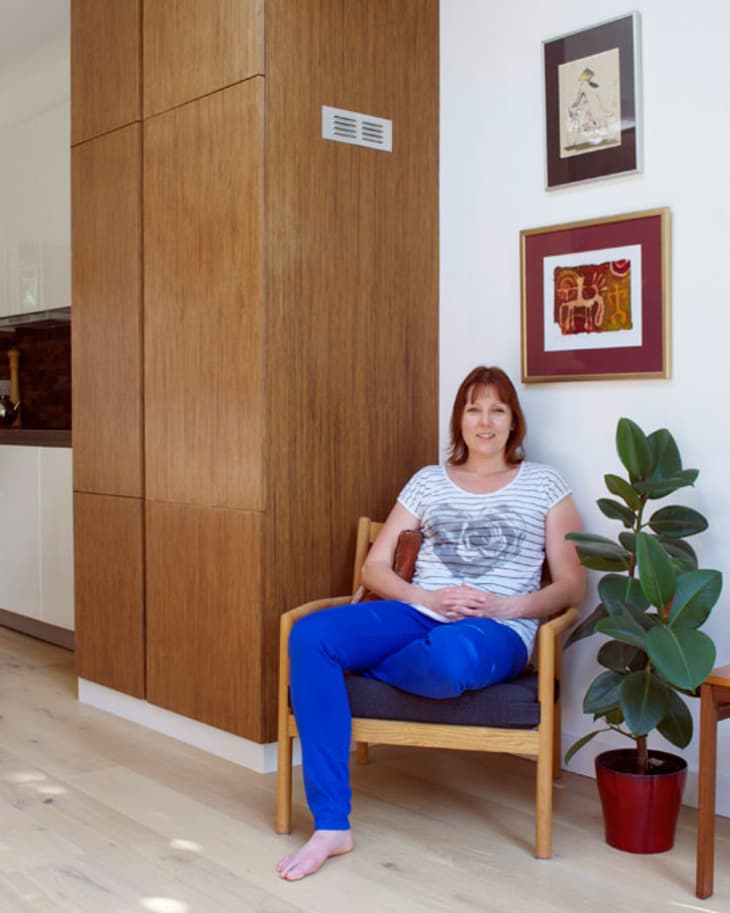 Woman sitting on a wooden chair in a modern room with wooden cabinets, framed art, and a potted plant.