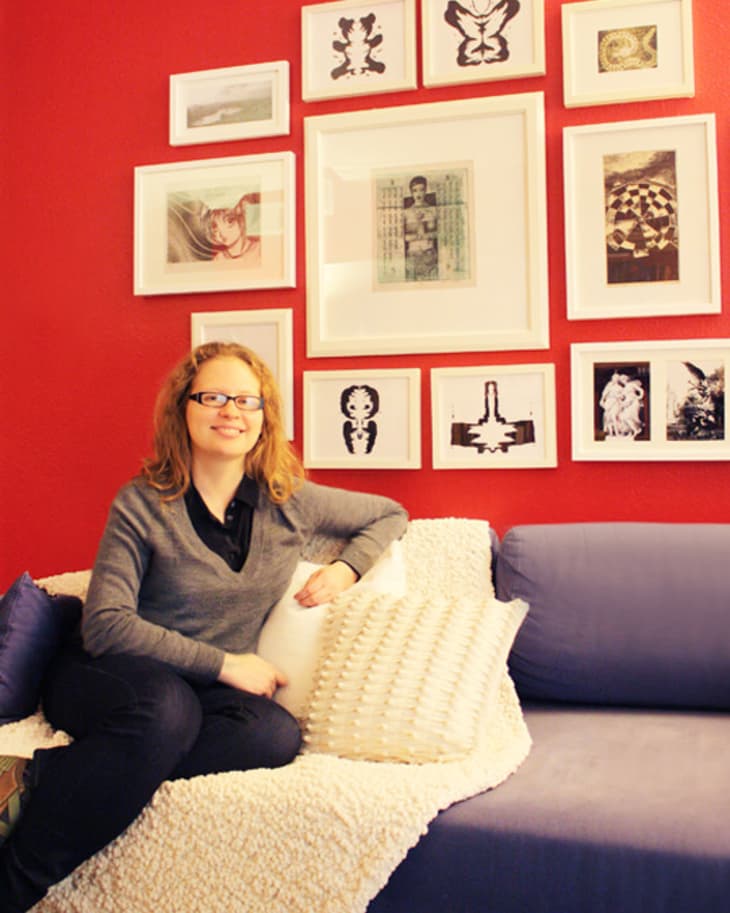 Woman sitting on a blue sofa with white pillows, against a red wall adorned with framed art.
