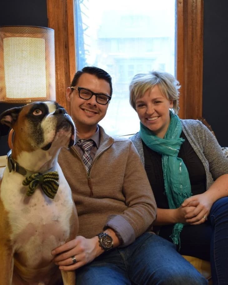 Couple sitting on a sofa with a large dog wearing a bow tie, next to a window with a lamp.