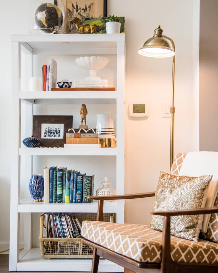 White bookshelf with books, globe, and decor items next to a patterned armchair and brass floor lamp.