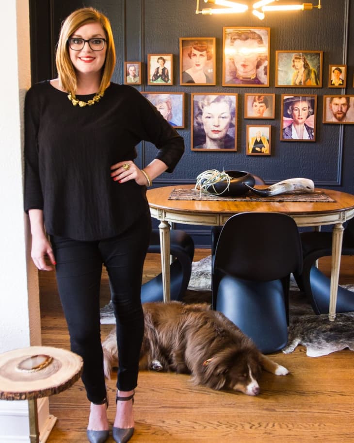 Woman in black outfit standing in a dining room with a gallery wall, modern chandelier, and a dog lying on the floor.