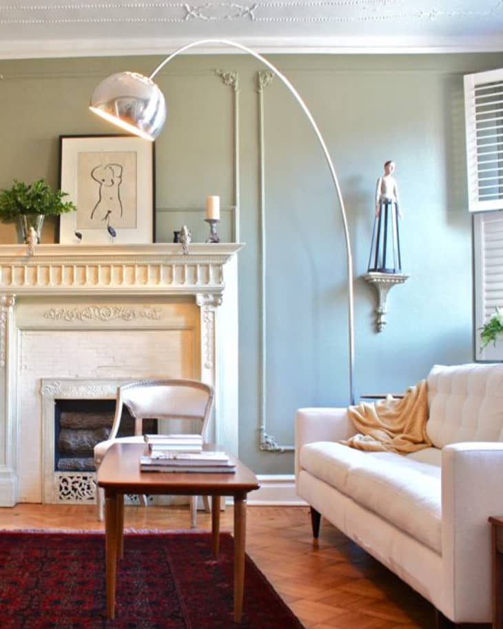 Elegant living room with a white sofa, arched floor lamp, ornate fireplace, and artwork on a light green wall.