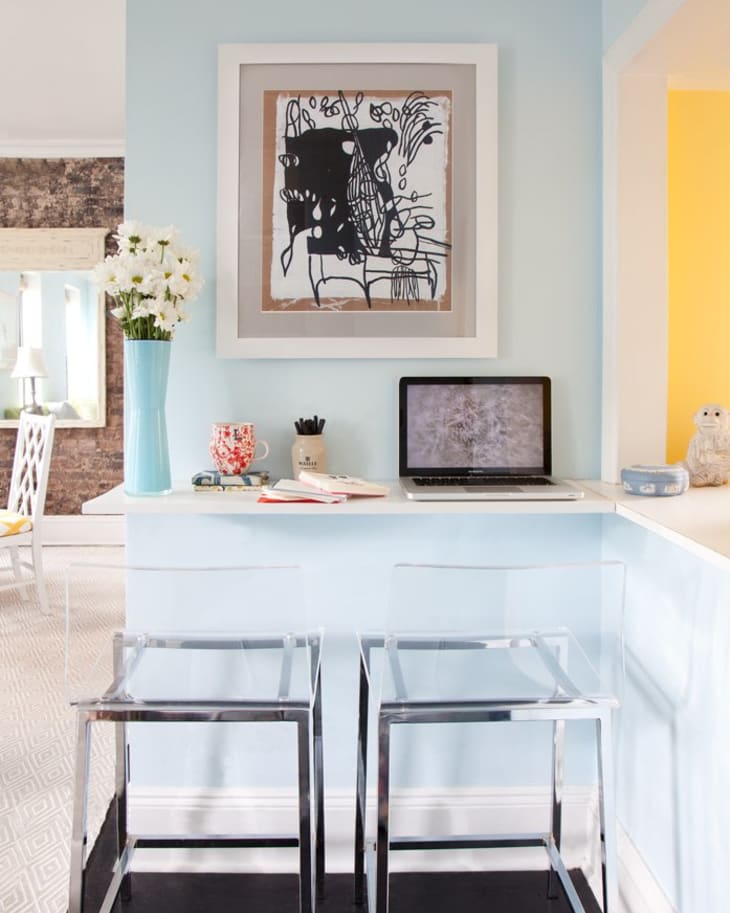 Home office nook with abstract art, laptop, clear stools, and blue walls.