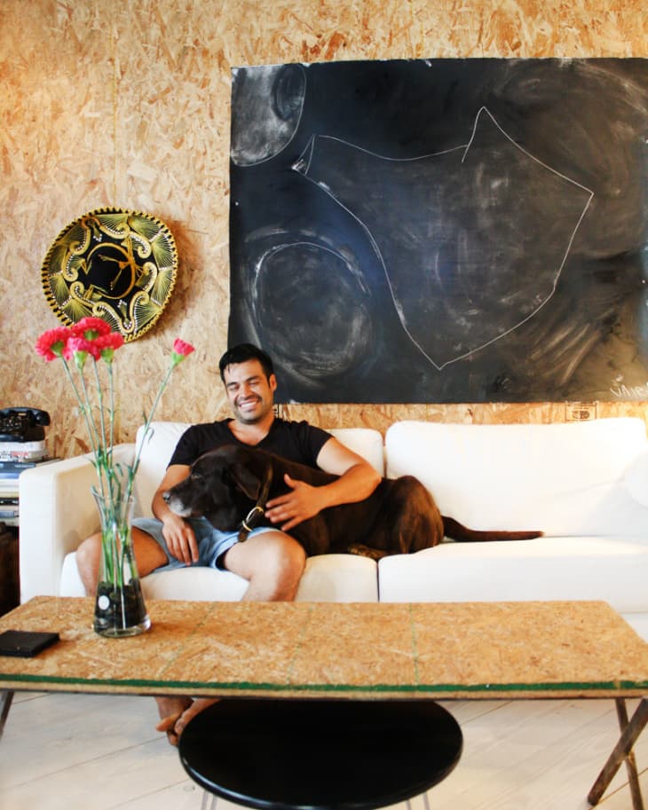 Man sitting on a white sofa with a dog, in front of a chalkboard and decorative plate, with flowers on a table.