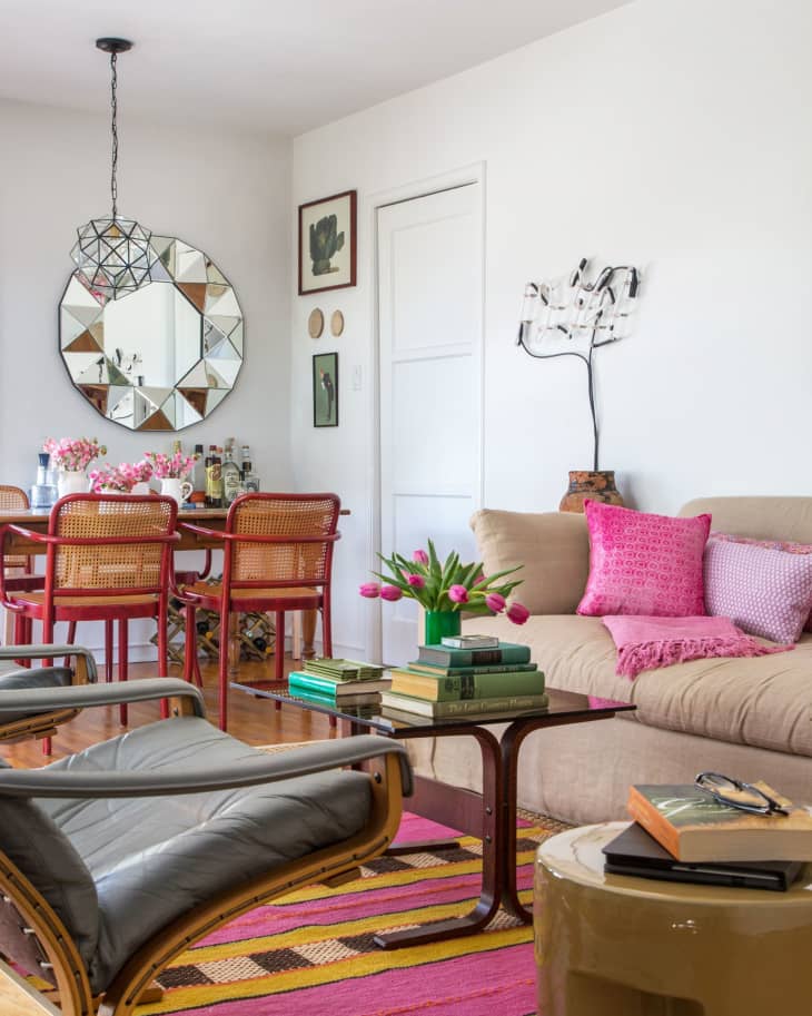 Living room with beige sofa, pink cushions, tulips on table, geometric mirror, and dining area with red chairs.