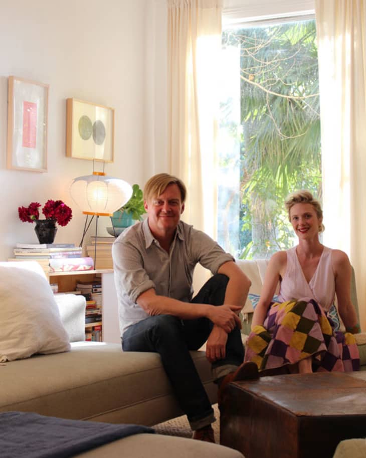 Two people sitting in a cozy living room with a beige sofa, wooden table, and large window with garden view.