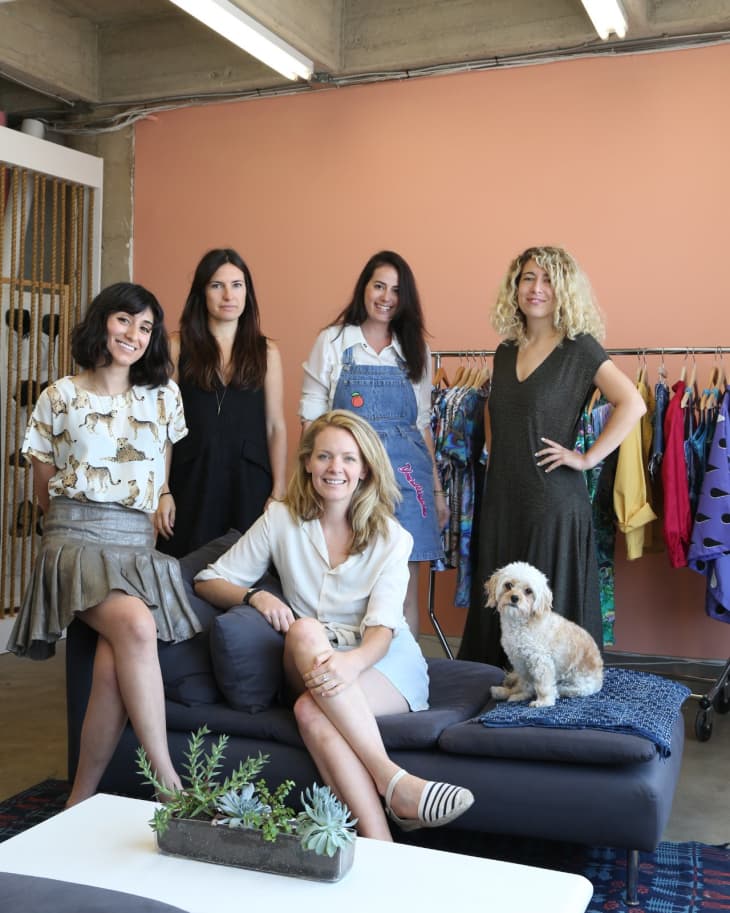 Five women and a dog in a room with a peach wall, clothing rack, and dark sofa with succulents on a table.
