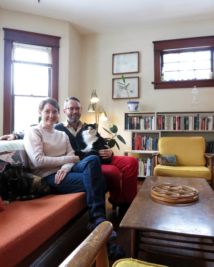 Couple sitting on an orange sofa with two cats, surrounded by bookshelves and yellow armchairs in a cozy living room.