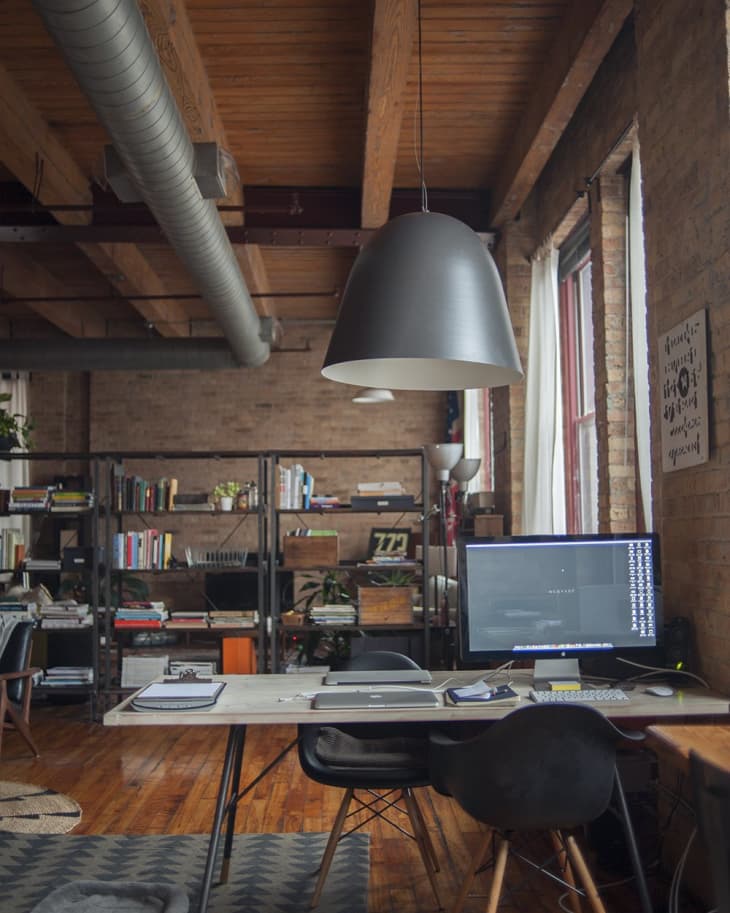 Loft office with wooden floors, large desk, computer, black chair, bookshelves, and industrial ceiling with exposed ductwork.