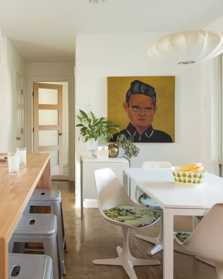 Dining area with white table, modern chairs, plant, and portrait painting on yellow background.