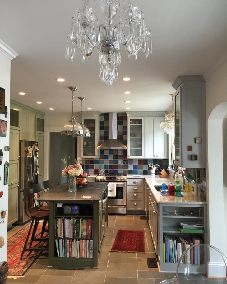 Kitchen with crystal chandelier, island with books, colorful tile backsplash, and stainless steel appliances.