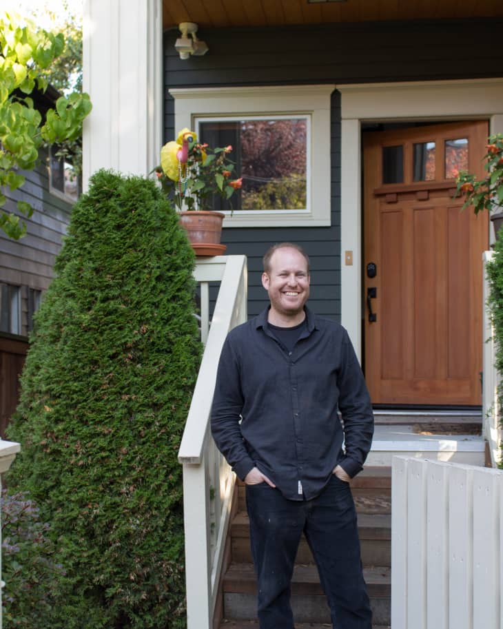 Man in black shirt standing on porch steps of a house with a wooden door, potted plants, and a large shrub.