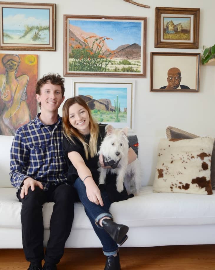 Couple sitting on a white sofa with a small dog, surrounded by eclectic framed artwork on the wall.