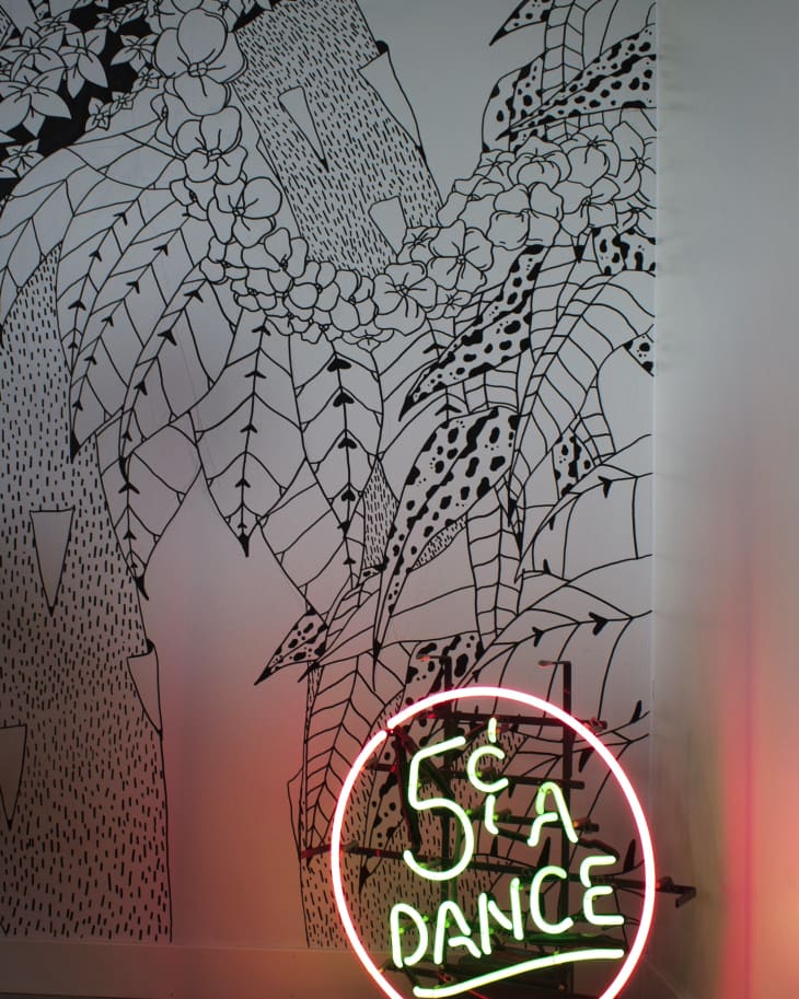 Neon sign reading "5¢ a Dance" in front of a black and white floral mural.