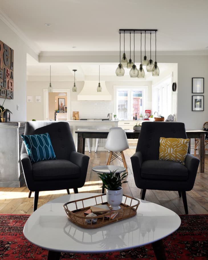 Living room with two gray armchairs, colorful cushions, white coffee table, and hanging pendant lights leading to a kitchen.