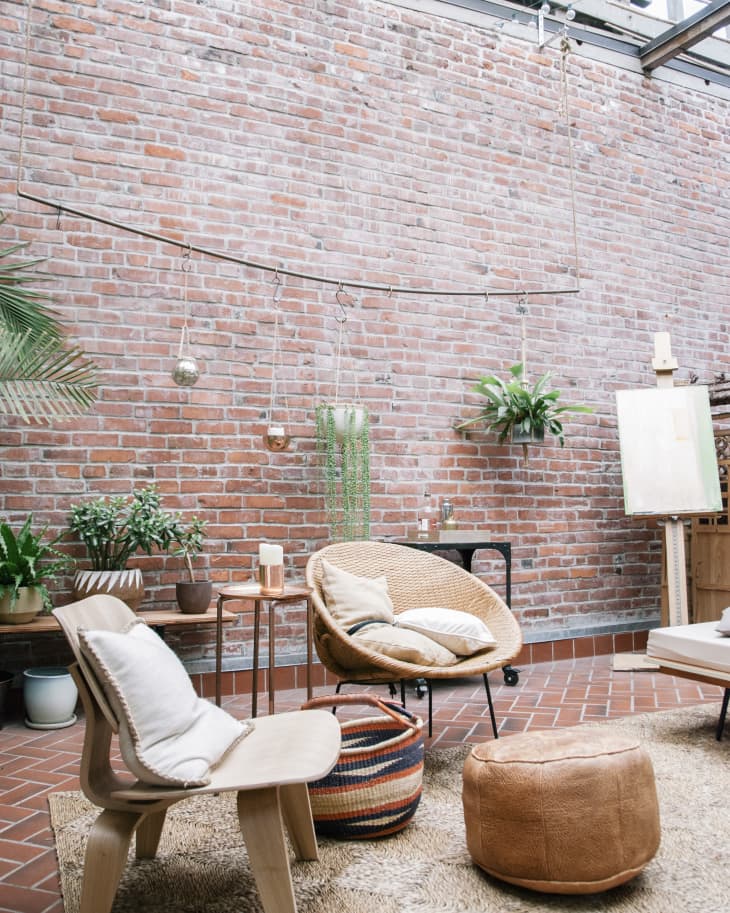 Cozy sunroom with brick walls, wicker chairs, potted plants, and a woven pouf on a textured rug.