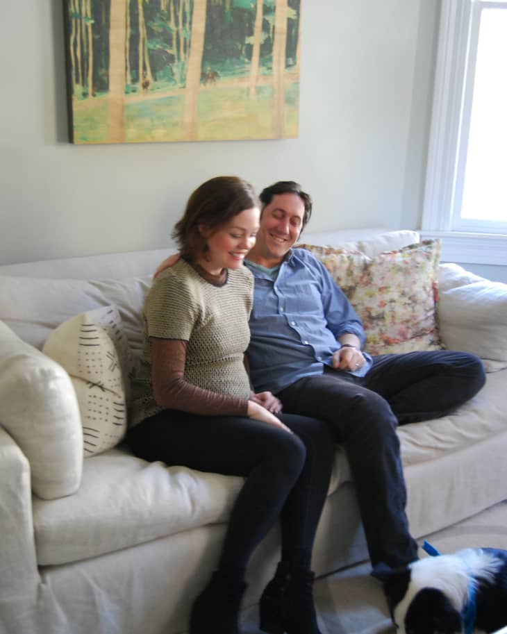 Couple sitting on a beige sofa with a dog on the floor, forest painting on the wall.